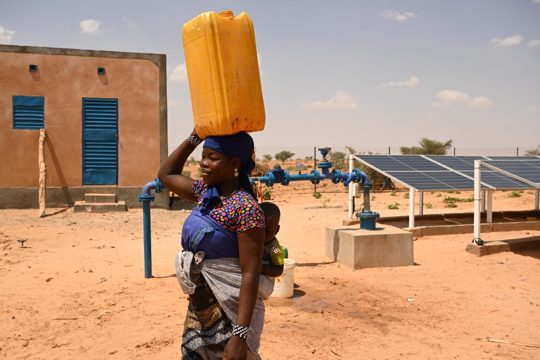 A woman in Africa, carrying a baby on her back, and holding a yellow water container on her head